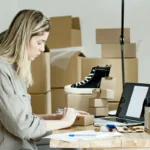 Wanita mengemas paket untuk toko online dikelilingi material kemasan dan laptop